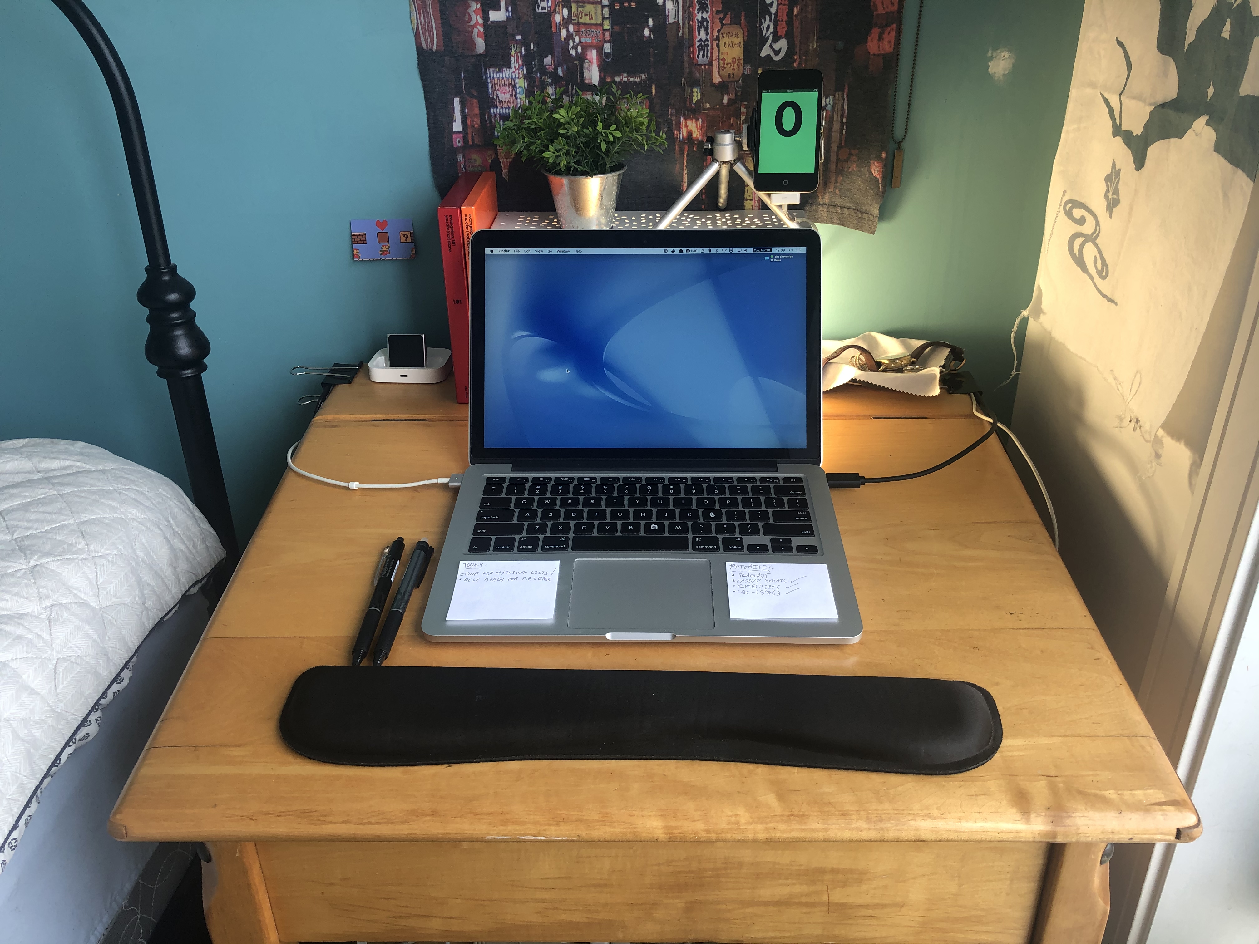 A laptop sits on a traditional wooden school desk. In front of it is a wrist rest, and to the right is an iPod Touch mounted on a small metal tripod. The laptop has two small pieces of paper on it either side of the trackpad, and two pens placed to its left.