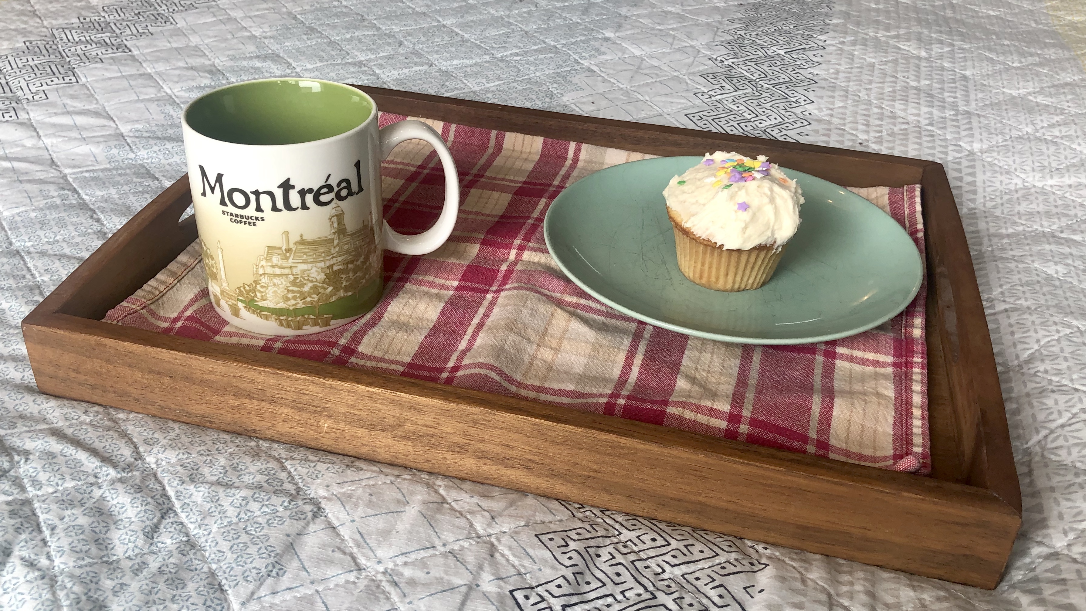 A small wooden lap tray with a mug on it, and a plate with a cup cake. The mug reads &ldquo;Montreal&rdquo; and the tray has a red patterned tea-towel underneath the mug and plate.